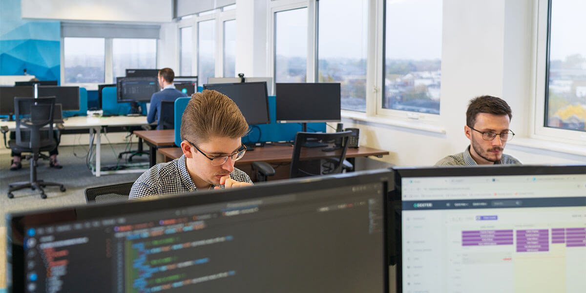 A camera shot over the top of two computer monitors with the team working in the background.