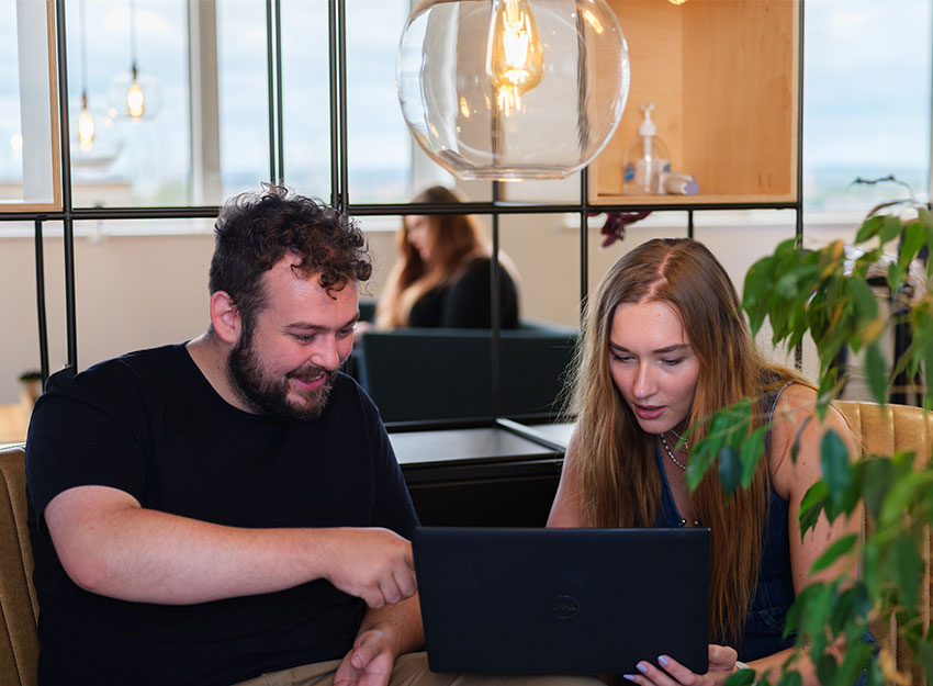 Two people sat in armchairs holding a laptop discussing work together. They are sat in a bright, large office with natural lighting from the windows behind.