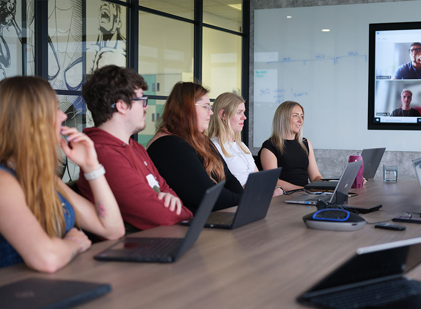 In a modern office people are sat in a meeting, each with laptops in front of them. At the end of the long table, a screen with a video call is in progress.