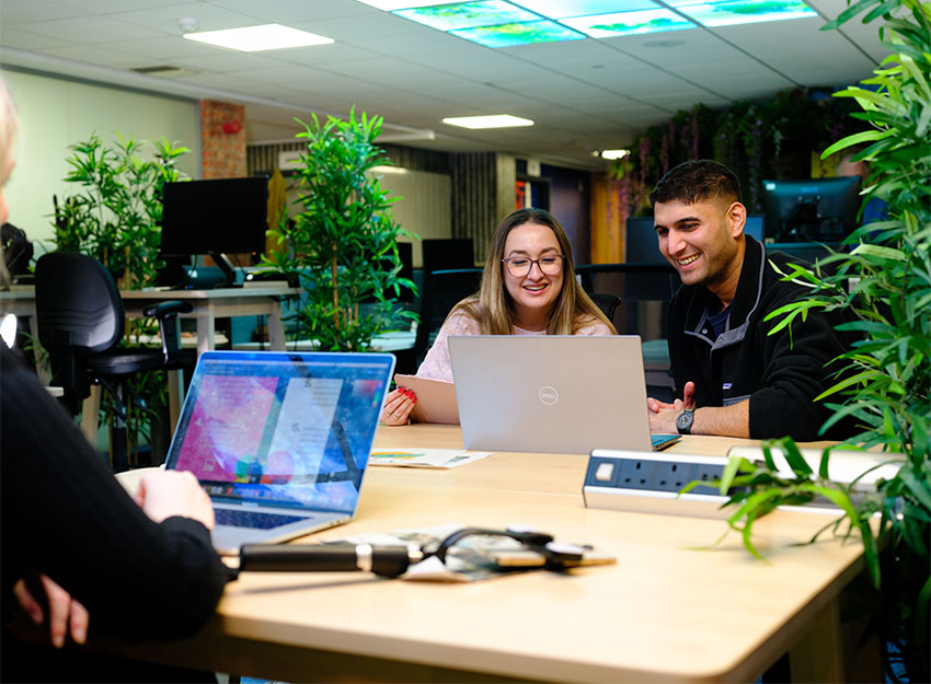 Three people are sat at a square table with laptops. A tech device for phone filming is placed on the table. Two people laugh as they discuss ideas.