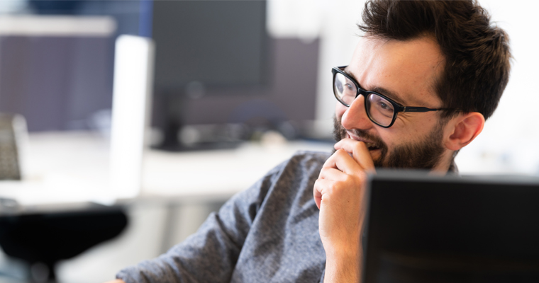 Tim is at his desk and has his hand to his mouth to stifle a laugh. He is wearing a grey sweater and thick black frame glasses