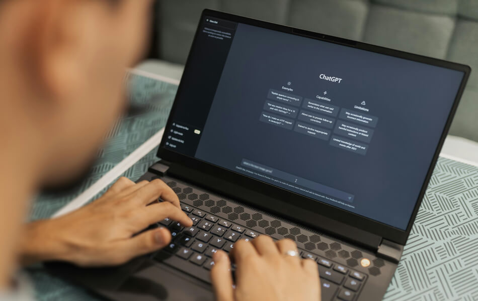 a person sitting at a blue textured table looking at their laptop whilst typing a prompt into OpenAI's ChatGPT generator