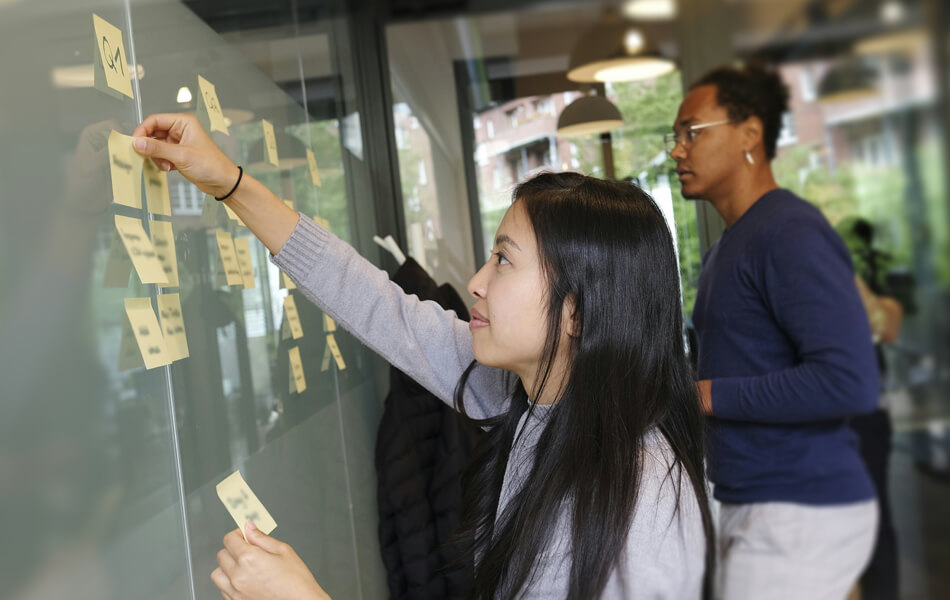 two work colleagues creating a campaign timeline by placing sticky notes on a glossy whiteboard inside a conference room