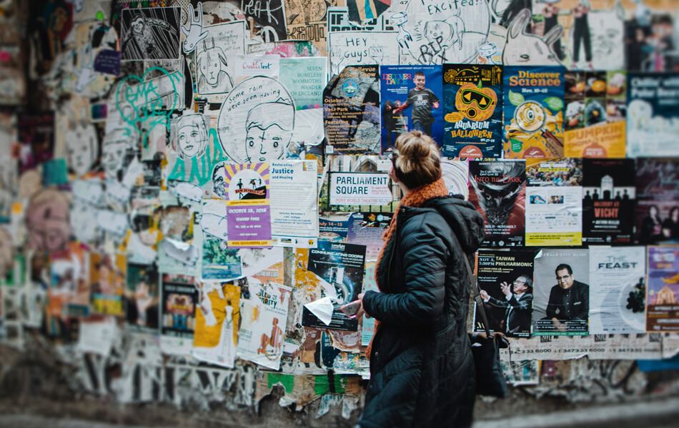 a young woman walking past a brick wall plastered with an array of peeling posters and adverts